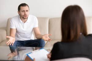 Young angry man sharing his problems with female psychotherapist, telling about troubles, expressing his feelings while female psychologist making some notes or filling in his personal information