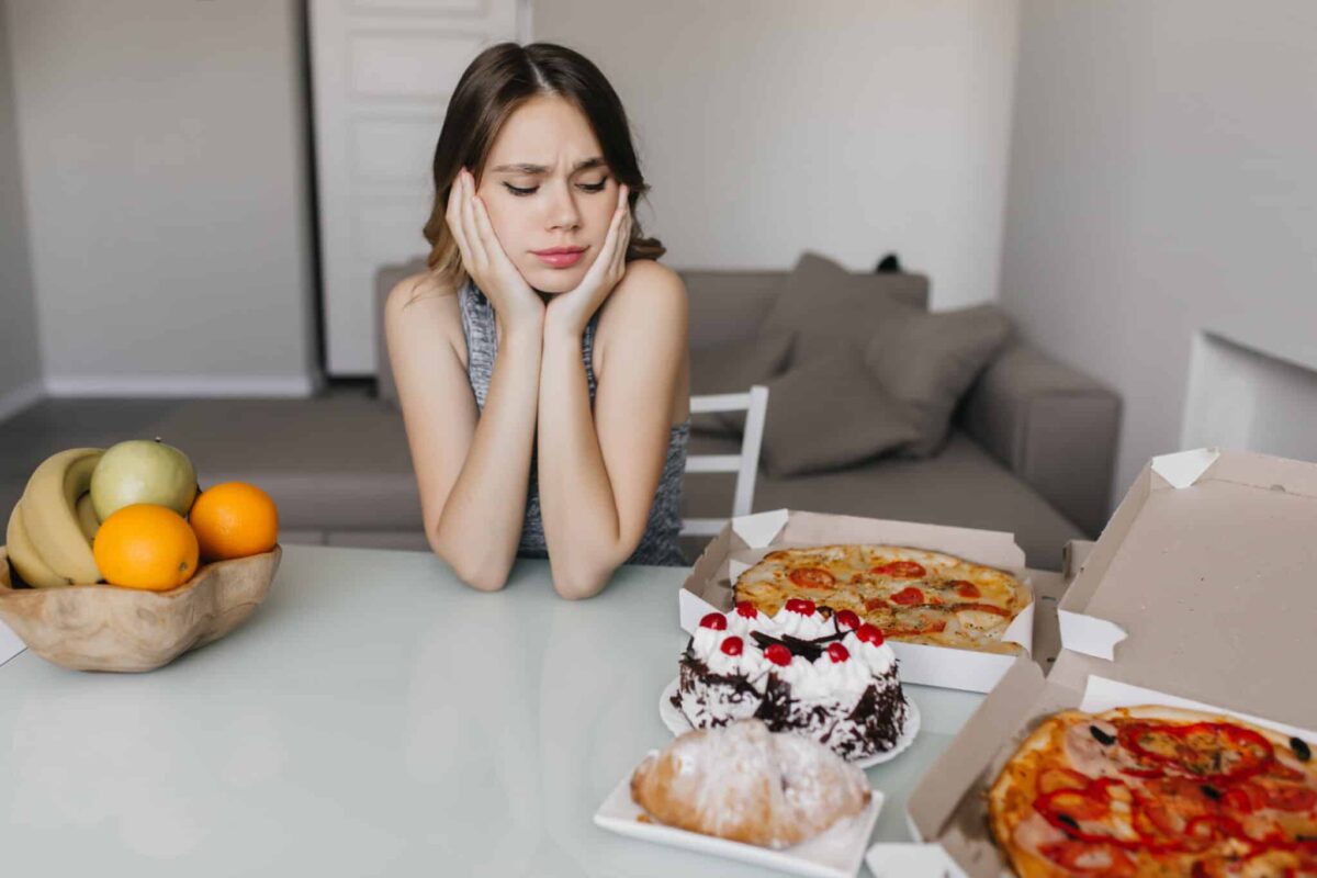 Sad curly woman looking at cake during diet. Blonde gorgeous female model posing with fruits and pizza, eating and disordered-eating behaviors massachusetts
