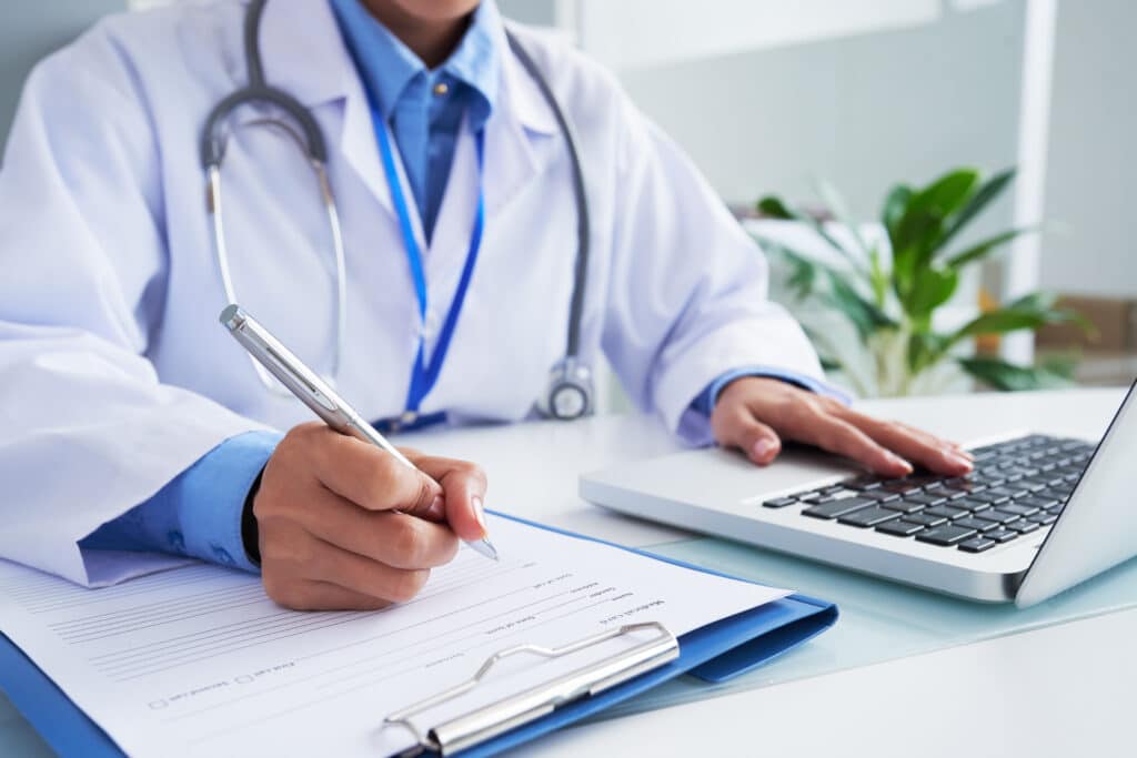 Doctor documenting patient information on a form while using a laptop, representing medical billing and coding in a clinical healthcare setting
